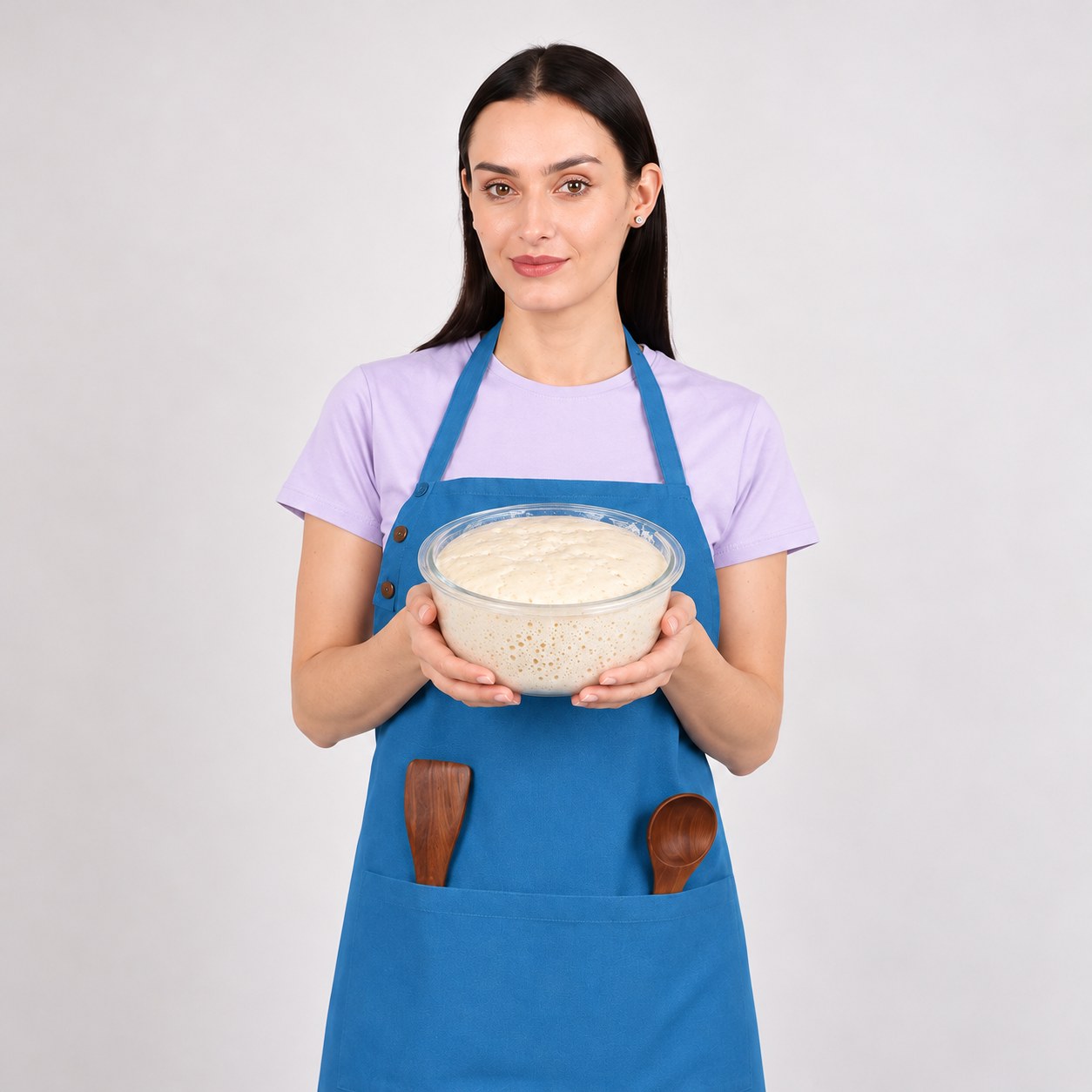 Emma in a blue apron holding a glass bowl of active sourdough starter, with wooden utensils in her pocket