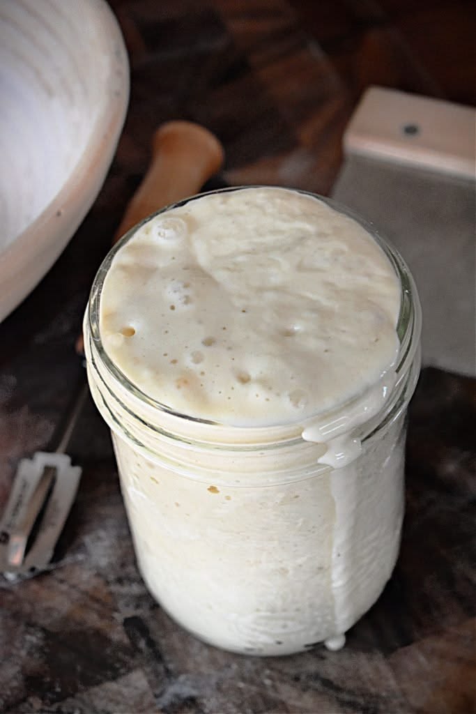Glass jar with active sourdough starter spilling over, next to a banneton