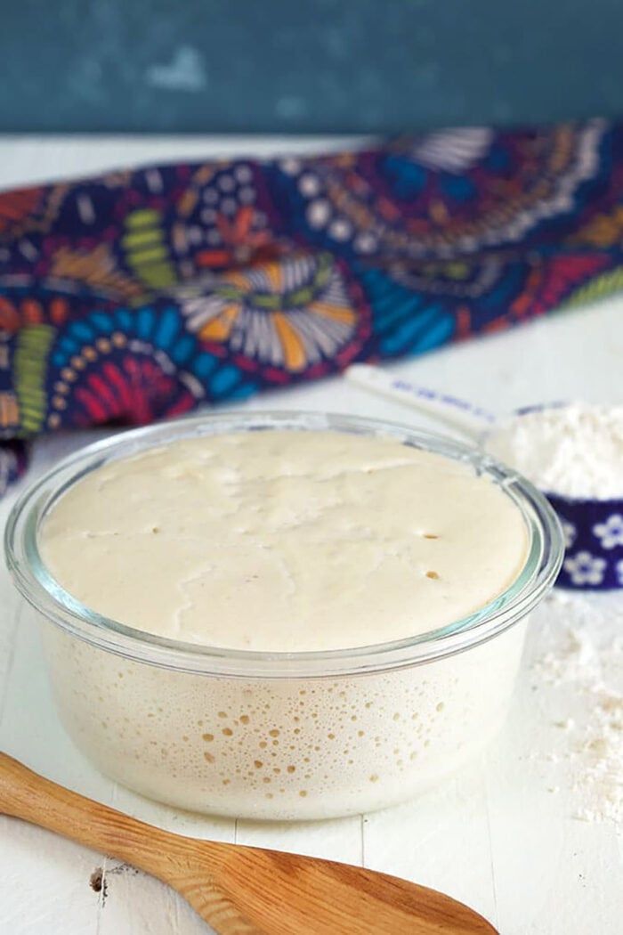 Glass bowl of active, bubbly sourdough starter next to a wooden spoon and flour